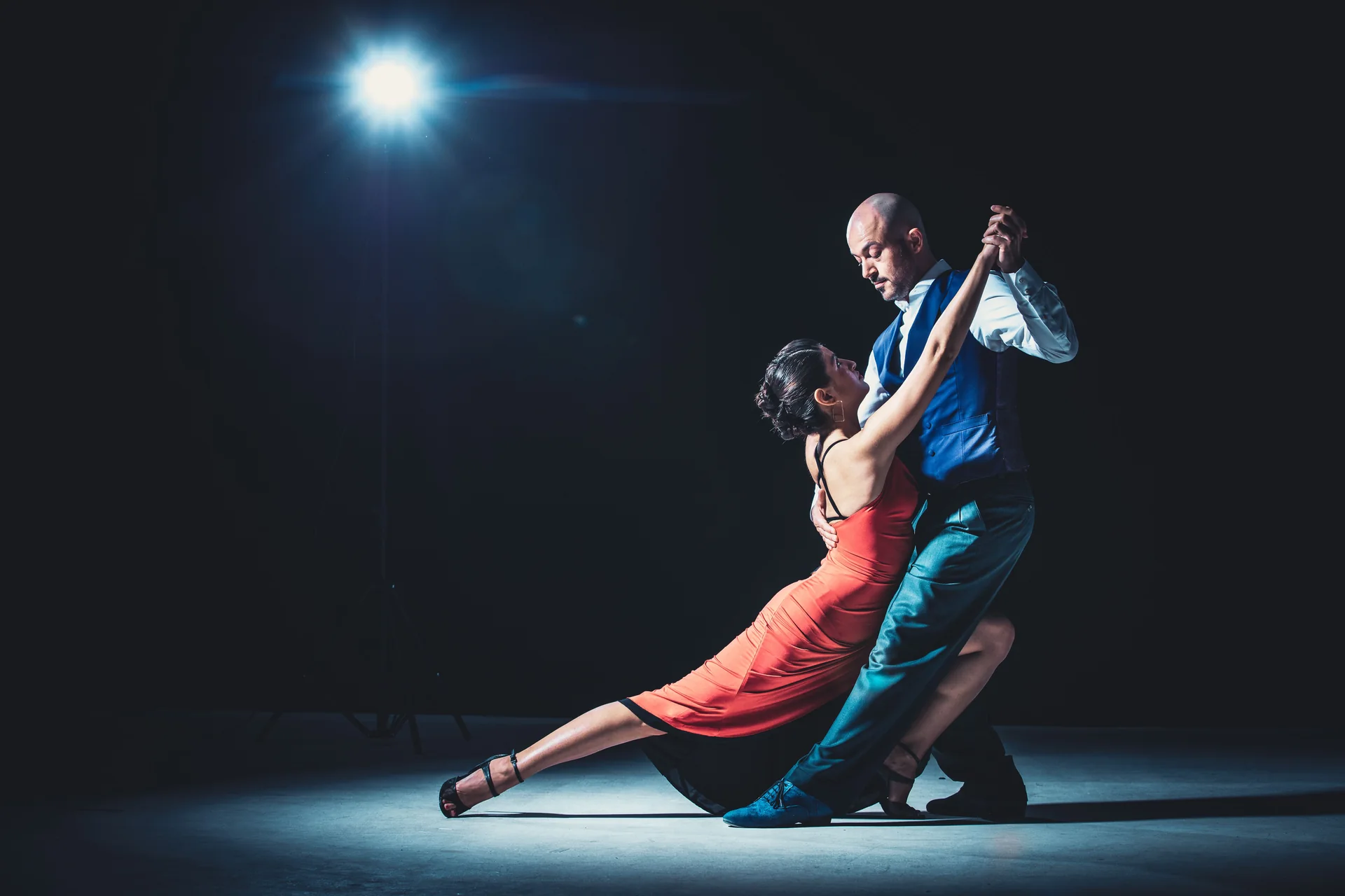 A tango couple in mid-dip under a single spotlight on a dark stage, the man in a waistcoat and the woman in a red dress (representative photograph of a tango stage performance, not Señor Tango's own room)